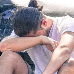 man sitting on the ground with his head in his arms in front of his wrecked vehicle