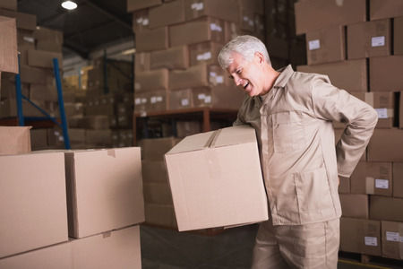 Older worker lifting a box in a warehouse and holding his back because he used improper lifting techniques.