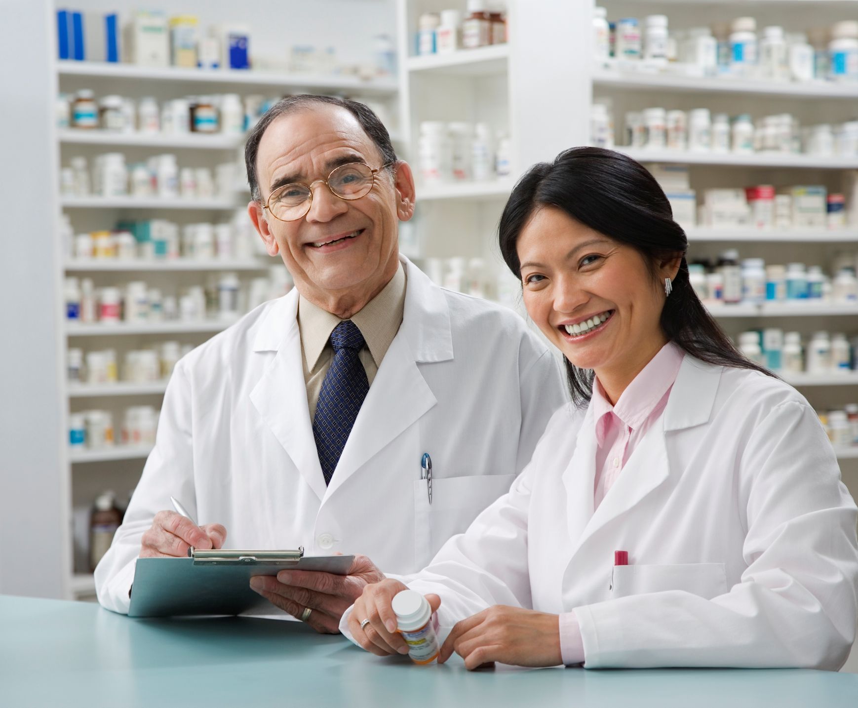 Two pharmacists at the counter and looking at the camera with shelves or medications behind them.