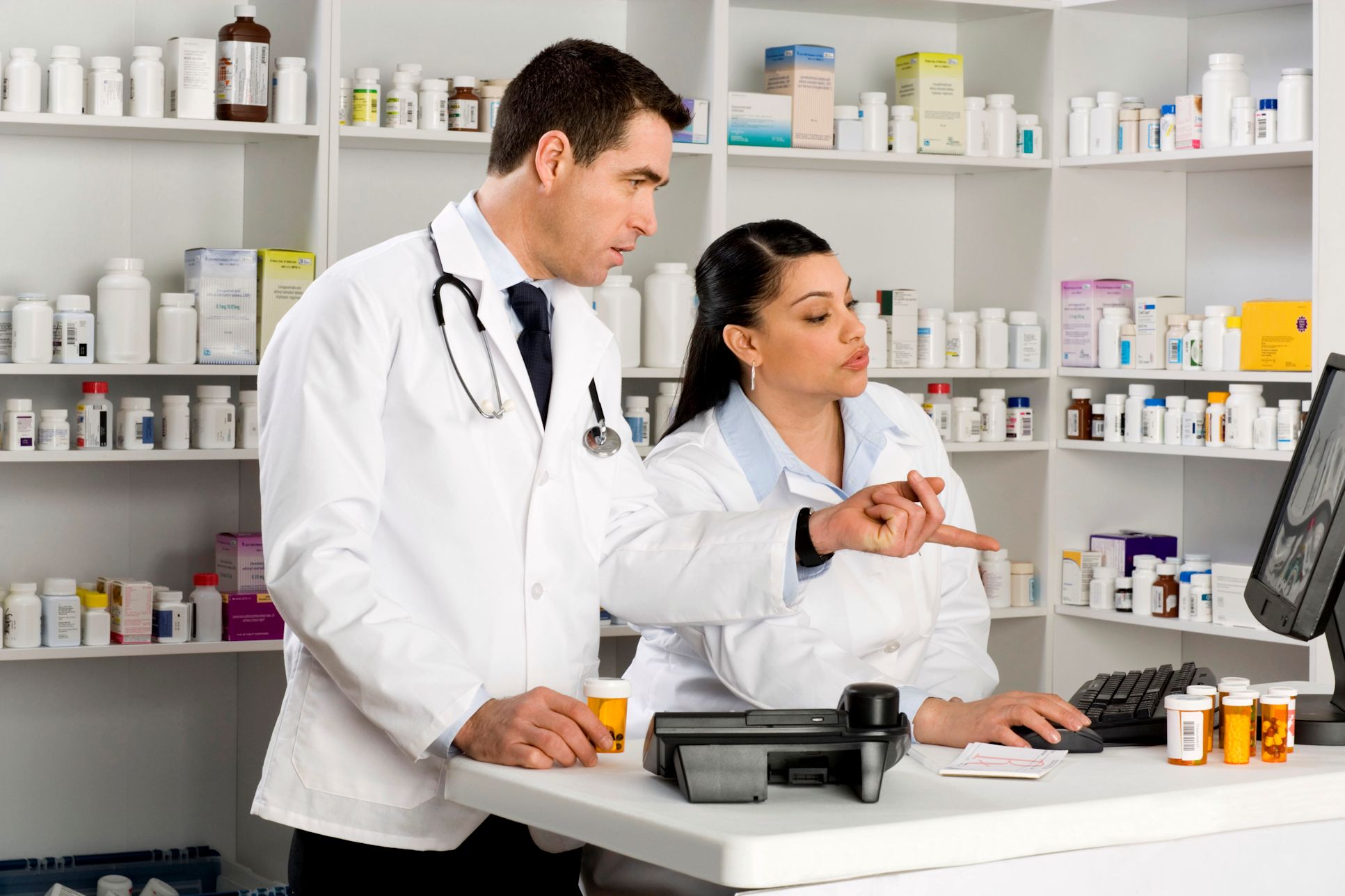 Pharmacist and tech looking at a computer monitor in a pharmacy. The wall behind them is filled with shelves of medication