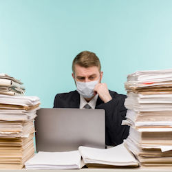 Employee wearing a mask and working at a desk in front of a laptop computer and the desk is stacked with papers.