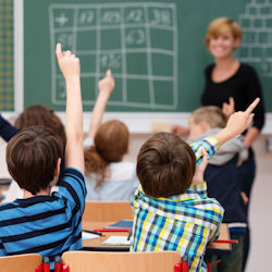 Teacher stands in front of class infront of a chalkboard. Students in the class have their hands raised.