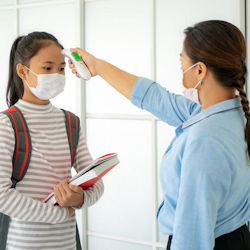 Adult, who has her back turned to the camera, takes the temperature of a student using a forehead therometer. Both people are wearing masks.