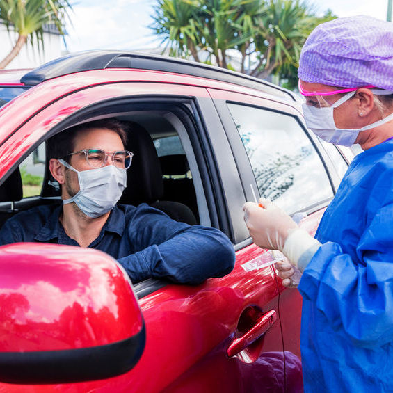 A man, wearing a mask, sits in a red car waiting for a COVID-19 test. There is a nurse in full protective gear standing outside the car.