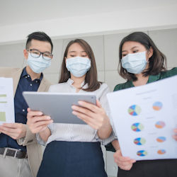 A man and two women wearing face masks look at a tablet. The two people on either side are holding reports.