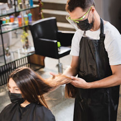 A stylist is looking at a womans hair. The woman has a gown
and face mask on. The stylist is wearing an apron, face mask, and face shield.