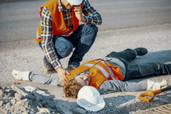 Man in an orange vest and hard hat kneels over another man laying on the ground. The man kneeling is on the phone calling for an ambulance.