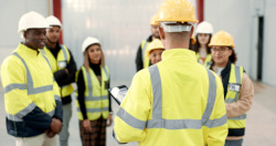 Back of man's head who is speaking to a group of people in front of him. Everyone is wearing yellow safety apparel and hard ahts