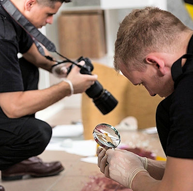 Two police officers investigating accident scene with a magnifying class and camera.