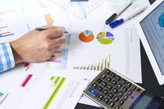 Worker at desk with computer and calculator with multiple graphs and charts laying on desktop