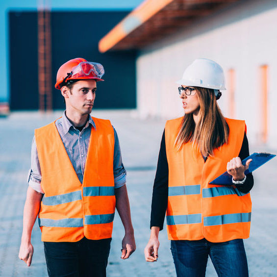 Two workers walking together both with helmets and safety vests one is holding a notebook