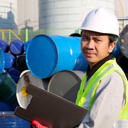 worker with clipboard evaluating the workplace. There is a stack of sealed drums on their side in the background.