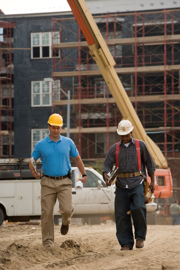 two men one contractor one construction worker outside walking through dirt with a building and crane behind them.