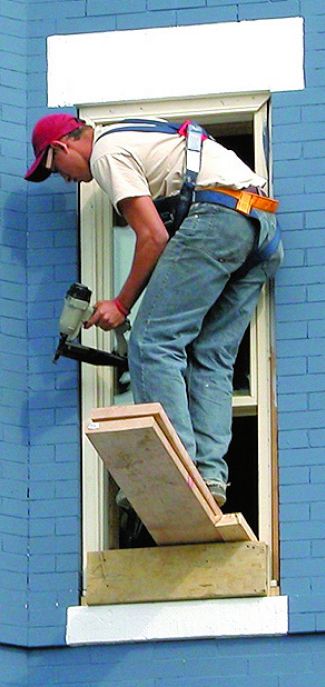 Worker standing on a boad sticking out of a window, holding a nailgun, and wearing a fall harness.